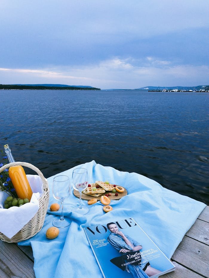 Enjoy a peaceful lakeside picnic with wicker basket, fruits, waffles, wine glasses, and a fashion magazine on a pier.