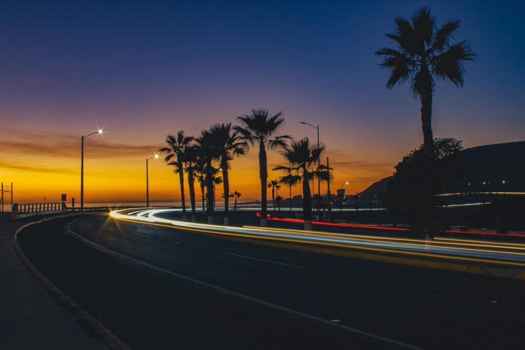pexels-photo-14086710-14086710 Stunning long exposure of Ensenada's coastal road at sunset, showcasing palm trees and light trails.