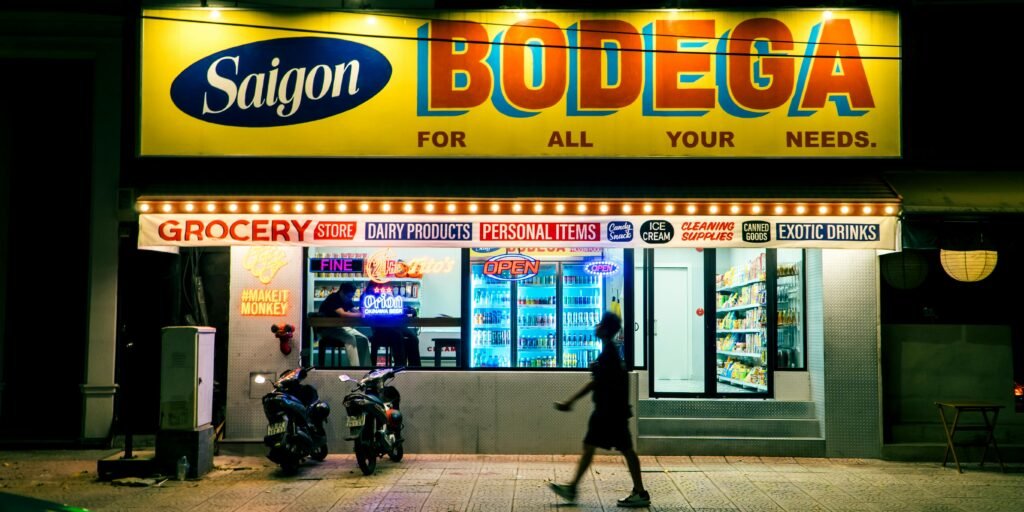 Colorful storefront of Saigon Bodega at night with a pedestrian silhouette and parked scooters.